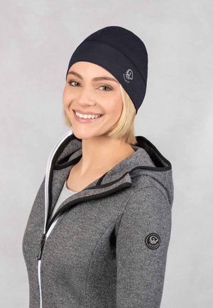 Granatowa czapka beanie wykonana z miękkiego, elastycznego materiału, z płaskim szwem i logo po boku. Noszona z szarym dresowym kapturem na zamek.