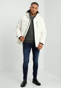 White padded jacket with a hood, front pockets, and a black lining, paired with a gray knit sweater and dark blue jeans. Black shoes.