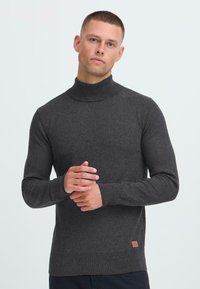 Giovane uomo con capelli corti che indossa un maglione a collo alto grigio scuro, in piedi con le mani delicatamente unite davanti a uno sfondo semplice.