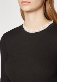 Svart ribbestrikket topp med lange ermer, utstyrt med en delikat hvit volangblondekrage som fremhever utringningen.