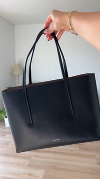 Saco tote de couro preto com alças longas, textura suave e um brilho sutil. Apresenta um detalhe de logotipo dourado na parte inferior.