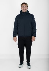 Doudoune bleu marine avec fermeture éclair sur le devant, design avec capuche et poignets élastiques, associée à un pantalon noir et des baskets blanches chunky.