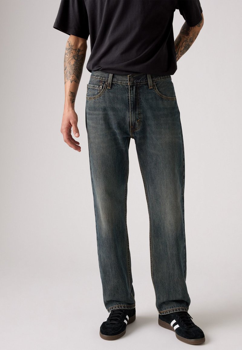 Vaqueros de denim azul con un corte relajado, que presentan áreas desgastadas y un diseño clásico de cinco bolsillos. Combinados con zapatillas negras y una camisa negra.