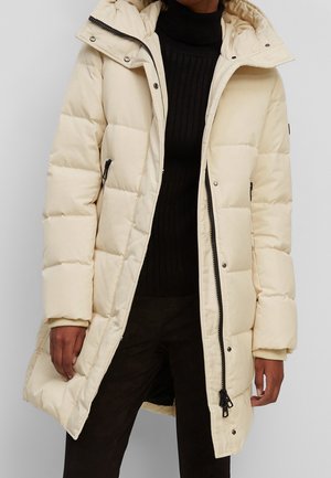 Persoon draagt een crèmekleurige lange gewatteerde winterjas met zwarte ritssluiting en drukknopen over een zwarte geribbelde coltrui.