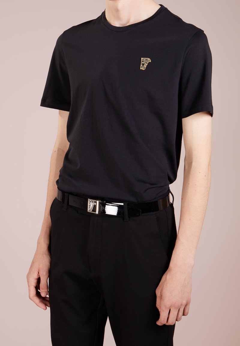 Mann trägt ein schwarzes T-Shirt mit einem kleinen goldenen Emblem auf der Brust, schwarze Hose und einen glänzenden schwarzen Gürtel mit metallischer Schnalle.