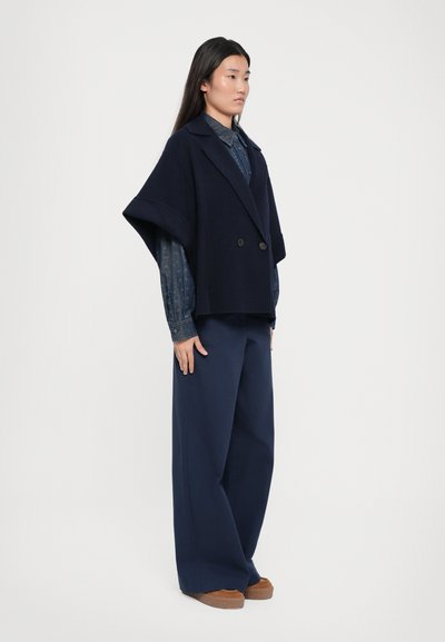 Marineblauwe wollen cape-achtige jas met brede mouwen en een dubbelzijdig ontwerp, gedragen over een gestreept denim overhemd en brede broeken.