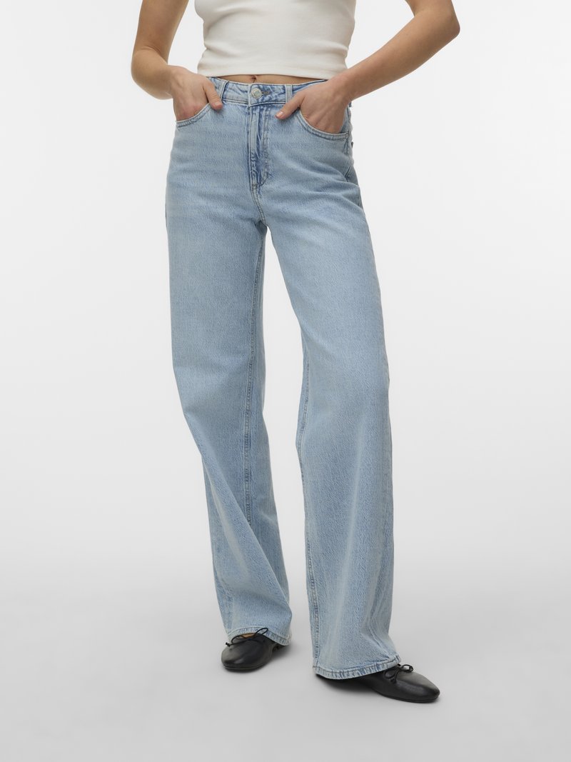 Osoba oblečená v svetlomodrých džínsach so širokými nohavicami, bielom krátkom topu a čiernych balerínach, stojaca s rukami v kapse.