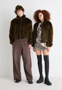 Dva modela nose umjetne krznene jakne u smeđoj i crnoj boji, uzorkovanu haljinu, oversized hlače i čizme do butina u crnoj boji. Teksturirani materijali i raznoliki oblici.