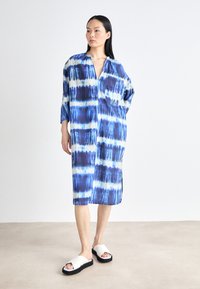 Rochie albă și albastră tie-dye, cu o croială lejeră, mâneci lungi, decolteu în V și o buzunară frontală, asortată cu sandale albe cu talpă plată.