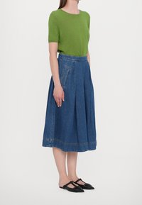 Maglione verde a maniche corte, gonna midi plissettata in denim blu, con tasche laterali, e scarpe flat nere con una strap texturizzata. Design semplice e classico.