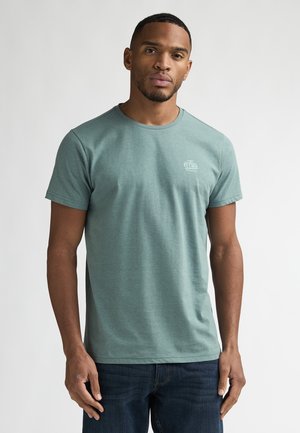 Tricou verde din bumbac cu mâneci scurte și decolteu rotund, cu un logo mic în partea stângă a pieptului, asortat cu blugi din denim închis la culoare.