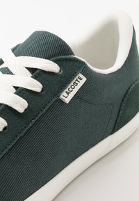 Baskets en toile vert foncé avec des lacets blancs et une semelle en caoutchouc. Présente une petite étiquette blanche Lacoste sur le côté. Texture lisse.