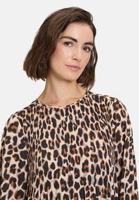 Bluusi, jossa on leopardi-kuvio ja pleatattu rakenne, sisältää mustia, ruskeita ja vaaleanruskeita värejä. Pyöreä kaula-aukko ja pitkät hihat, joissa on pehmeä tuulen vaikutus.