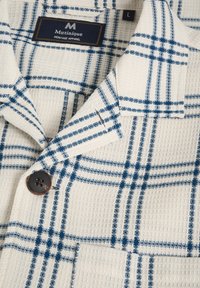 Fehér, textúrált szövet póló kék kockás mintával, fekete gombbal és "Matinique Heritage Apparel" feliratú címkével.