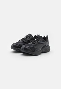 Paire de baskets de sport noires avec dessus en mesh et synthétique, fermetures à lacets, et semelles en caoutchouc épaisses et texturées sur fond blanc.