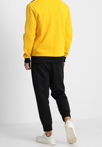 Gul fleece-sweater med svarta detaljer och ribbade muddar, ihop med svarta joggerbyxor och vita sneakers, sedd bakifrån.