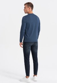 Langärmliges dunkelblaues Hemd mit Rundhalsausschnitt, kombiniert mit dunklen Jeans und weißen Sneakern. Einfaches, figurbetontes Design mit minimalen Details.
