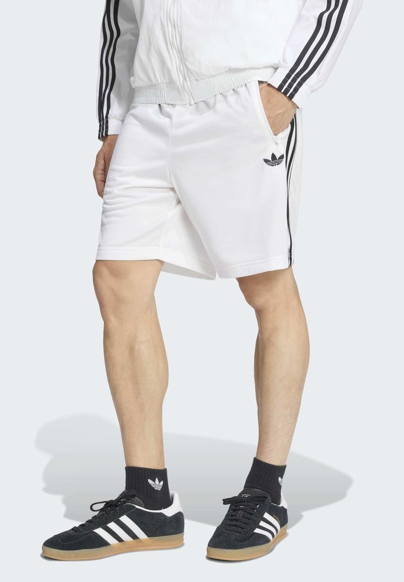 Persona che indossa pantaloncini e giacca Adidas bianchi con strisce nere, calzini neri Adidas e scarpe da ginnastica nere Adidas con strisce bianche.