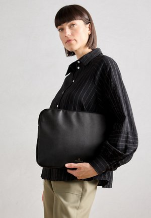 Black textured leather bag, rectangular shape, features a gold brand logo, held against a striped blouse with a collared design.