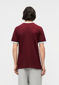 Άτομο με μεσαίου μήκους σκούρα μαλλιά, ορα τό από πίσω, που φοράει μπλούζα με κοντά μανίκια σε μπορντό χρώμα με ανοιχτό γκρι περιθώρια και γκρι παντελόνι.