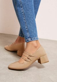 Béžové boty s blokovým podpatkem a špičatou špičkou, vybavené třemi úzkými pásky na kotníku s kovovými přezkami. Nošeny s modrými džínami.