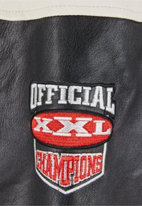 Patch en cuir noir avec texte brodé en blanc, rouge et noir : "OFFICIAL XXL CHAMPIONS" avec un logo bien visible en dessous.