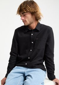 Chemise noire à manches longues en tissu doux, avec un col à boutons, une poche poitrine et des boutons blancs, associée à un jean bleu clair.