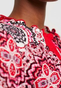Blusa de patrón floral multicolor que presenta tonos rojos, negros y rosas, con un escote con volantes y tela texturizada. Vista en primer plano del diseño.