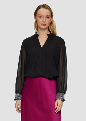 Schwarze Bluse mit gefaltetem Vorderteil, V-Ausschnitt und transparenten langen Ärmeln. Verfügt über gemusterte Manschetten. Kombiniert mit einem leuchtend rosa Satinrock.