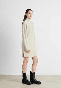 Abito lavorato a maglia beige con colletto alto, maniche lunghe e spacchi laterali; abbinato a stivali neri alla caviglia con suola spessa e dettaglio con fibbia.