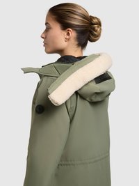 Olijfgroene jas met een capuchon gevoerd met nep bont, zwarte binnenkant en zwarte knoopdetails. Minimalistisch ontwerp, aan de voorkant opengezipt.