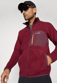 Man wearing a burgundy Columbia fleece jacket with zippered pockets and a black cap and dark sunglasses, standing against a plain background.