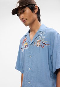 Ανοιχτό μπλε κοντομάνικο πουκάμισο με κεντημένα σχέδια, που απεικονίζουν ήλιο, κύματα και γραφή. Το ύφασμα έχει υφή με ελαφρώς ζαρωμένη εμφάνιση.