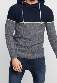 Granatowo-szara dzianinowa bluza z kapturem o teksturowanej tkaninie, ze sznurkami i białą linią akcentującą na wysokości klatki piersiowej. Niebieskie logo przy dolnym brzegu.