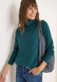 Maglione dolcevita teal con lavorazione a tricot e polsini a coste, abbinato a jeans blu casual. Una borsa grigia è appoggiata sulla spalla.