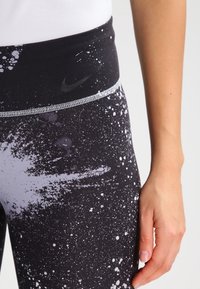 Leggings Nike avec une taille haute, ornés d'un motif éclaboussure de peinture noir et blanc, en tissu extensible, et avec un détail de logo réfléchissant.
