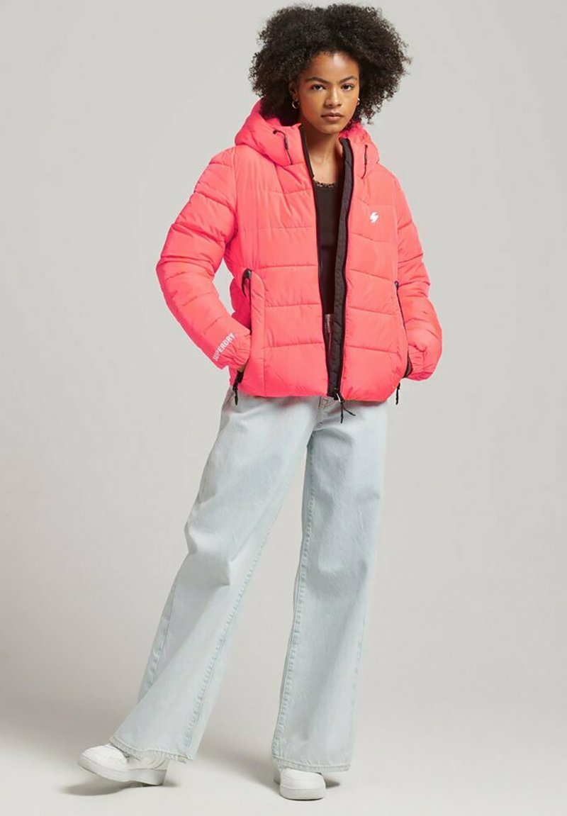 Giacca puffer rosa brillante con cappuccio, caratterizzata da un design trapuntato e una chiusura frontale con zip. Abbinata a jeans a gamba larga azzurri e sneakers bianche.