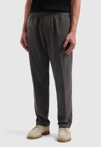 Man in een donkergrijs elastische taillebroek en beige veterschoenen, staand op een lichtgrijze achtergrond.