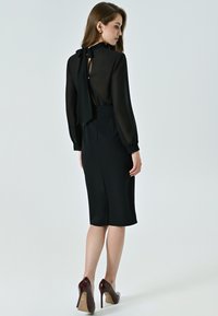 Schwarzes Kleid mit transparentem Rücken und Bindedetail, mit langen Ärmeln und figurnaher Silhouette. Getragen mit bordeauxfarbenen High Heels.