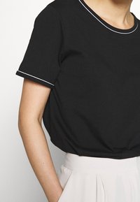 Schwarzes Kurzarmoberteil mit rundem Ausschnitt, ausgestattet mit weißen Einsätzen entlang des Kragens und der Ärmel. Weiches Material mit einer lockeren Passform.