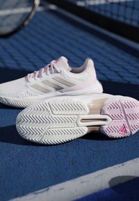 Par af hvide og lyserøde tennissko på blå bane, den ene viser sålens mønster, med tennisracket og net i baggrunden.