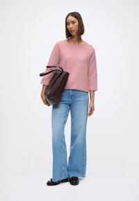 Κόκκινη και λευκή ριγέ μπλούζα με μακριά μανίκια, μπλε καμπάνα τζιν, κρατώντας μια υφασμάτινη μπορντώ τσάντα, μαύρα παπούτσια με στρογγυλή μύτη και περίπλοκο σχέδιο κορδονιών.