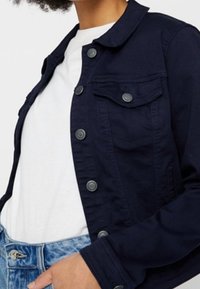 Giacca in denim blu navy con chiusura a bottoni, due tasche sul petto e colletto. Il materiale sembra robusto con una texture liscia.