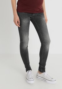 Osoba oblečená v vybledlých černých skinny džínách a bílých nízkých teniskách stojící proti jednobarevnému světlému pozadí.