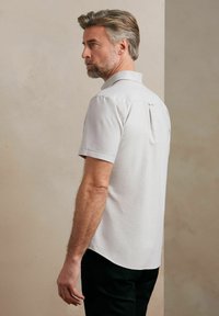Jasnoszara koszula krótko-sleeve z kołnierzykiem, z plisą z tyłu, zaokrąglonym dołem i gładką fakturą materiału.