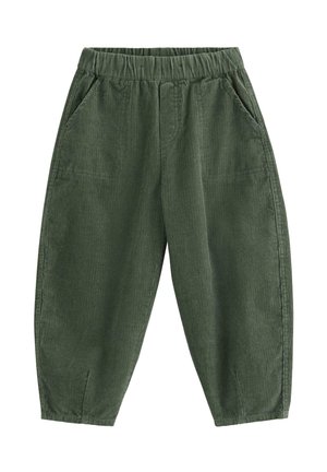 Junger Junge lächelt, trägt einen cremefarbenen Zopfpullover, lockere grüne Cordhosen und mehrfarbige Sneakers, steht mit Händen in den Taschen.