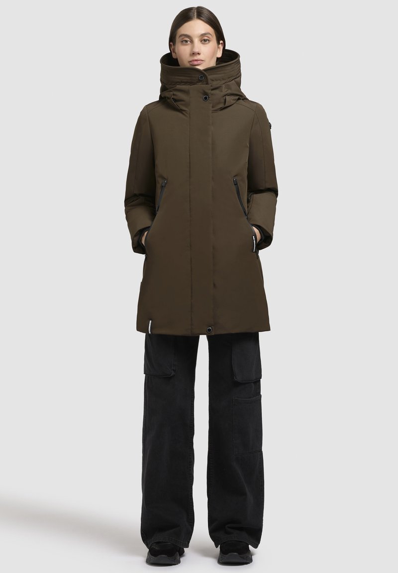 Abrigo acolchado de color verde oliva con cuello alto, bolsillos laterales con cremallera y un diseño recto. Combinado con pantalones cargo negros y zapatos.