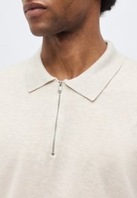 Mann trägt ein hellbeiges Hemd mit Kragen und einem vertikalen silbernen Reißverschluss, der am Ausschnitt teilweise geschlossen ist.