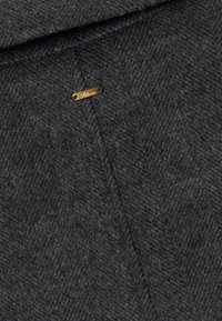 Un tissu en laine à chevrons gris foncé avec une petite étiquette en métal doré gravée avec "Billion" attachée près d'un bord replié.