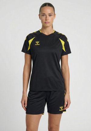 Vrouw staand in zwarte sportkleding met gele accenten, waaronder een T-shirt met korte mouwen en een korte broek, beide met Hummel-logo's.